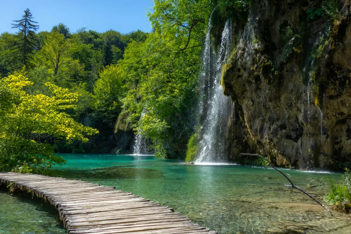 Wooden walkway crossing clear water beside waterfalls and forest at Plitvice Lakes